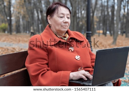 An elderly woman in a bright terracotta coat sits on a bench in an autumn Park with a laptop and selects goods online