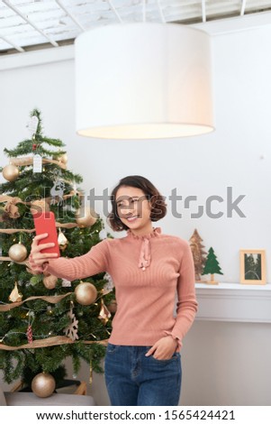 Funny asian girl taking selfie pictures on smartphone camera at home near Christmas tree