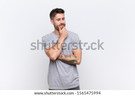 young handosme man smiling with a happy, confident expression with hand on chin, wondering and looking to the side against flat color wall
