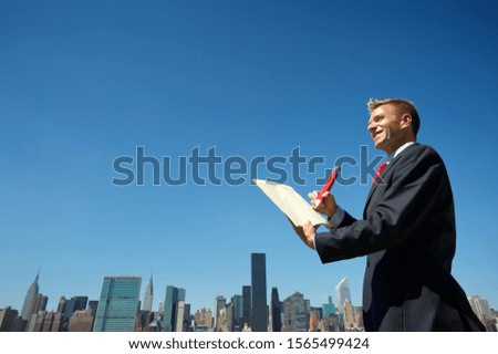 Smiling businessman standing at the city skyline with an oversize red pencil and notepad