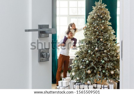 christmas morning, Dad and daughter wearing a Santa Claus opening a door,looking at the camera, behind them a Christmas tree decorated, with gifts in his foot. The sun gives cozy atmosphere