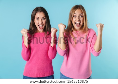 Photo of delighted caucasian women in pink sweaters screaming with winners gesture isolated over blue background