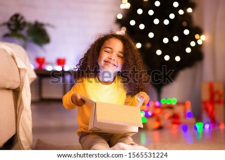 Christmas time. Cheerful afro child enjoying opening gift, sitting near Xmas tree, empty space