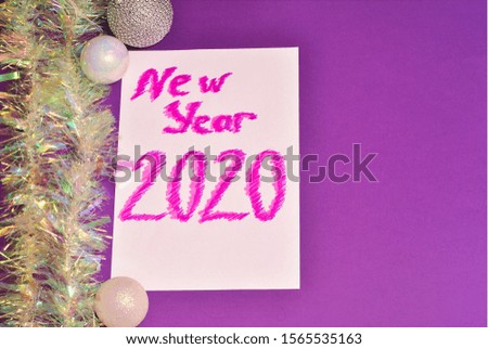 New year 2020 handwritten caption on white paper decorated with sparkle decorations on the purple background with colorful shiny tinsel and shiny silver white balls. Winter holidays celebration