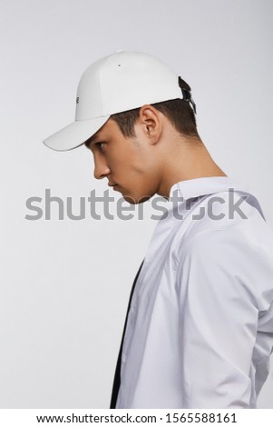 Cropped side photo of a dark-haired man, wearing white baseball cap with lettering "none", black t-shirt and white shirt. He is posing on grey background and looking straight. 