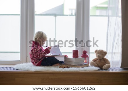 Sweet blonde child, boy, sitting on window shield with teddy bear friend toy, writing letter to Santa Claus and reading book with toy