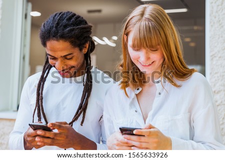 Stock photo of multicultural couple with mobile phone in hand and looking at the screens on the outside of a house