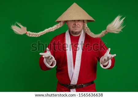 Male actor dressed as Santa Claus with long braids hairstyle and Asian hat posing on green chrome background