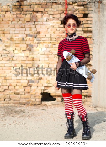Young alternative goth girl with piercing with white longboard on a brick wall background. Beautiful emo female model holding skateboard in hands. Alternative teen lifestyle