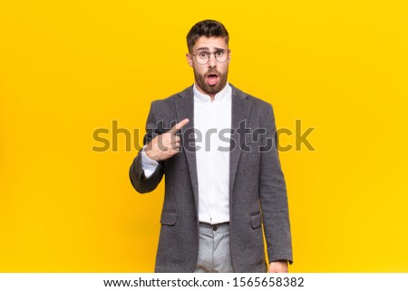 young handosme man looking shocked and surprised with mouth wide open, pointing to self against flat color wall
