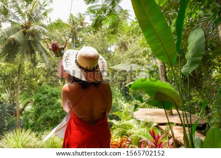Young lady in red dress taking a photo with a mobile phone while her friend swinging on a Bali swing in the middle of the rainforest jungle on this beautiful island in Indonesia  