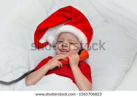 Little boy lying on white linen in the Santa hat laughing and holding a red handset.