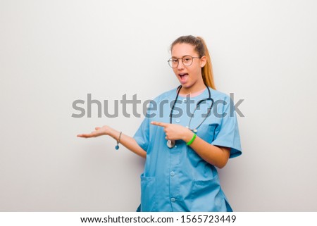 young latin nurse smiling, feeling happy, carefree and satisfied, pointing to concept or idea on copy space on the side against white wall