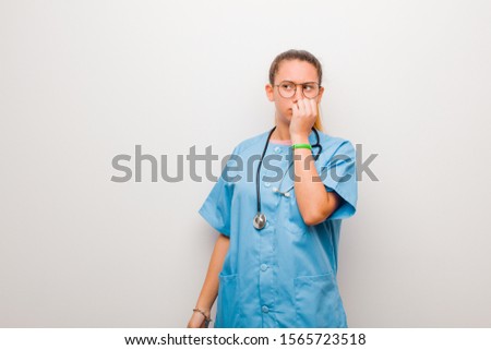 young latin nurse looking worried, anxious, stressed and afraid, biting fingernails and looking to lateral copy space against white wall
