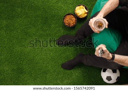 A man sits at the TV and watches a football match. TV remote. Soccer ball. View from above. Copy space.