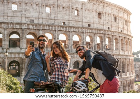 Three happy young friends tourists with bikes at Colosseum in Rome taking pictures and selfies with smartphone