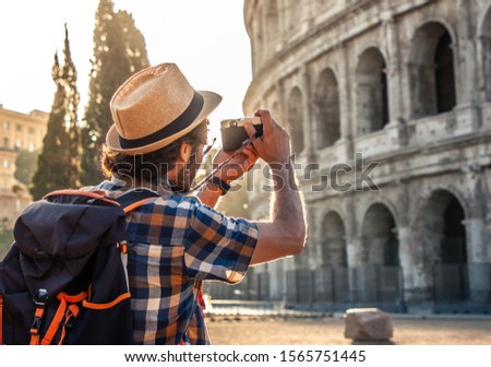 Happy young man tourist wearing shirt and hat taking pictures with vintage camera at colosseum in Rome, Italy at sunrise.