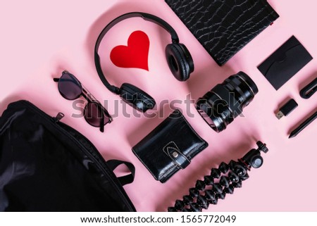 Trendy black accessories and contemporary gadgets on pink background, overhead view with copyspace. Modern headphones with heart symbol, camera lens, wallet, sunglasses, notebook and purse, flat lay