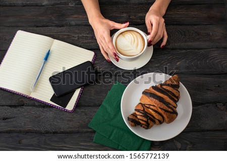 Woman drinks cappuccino in a French cafe. Croissant, notapad, smartphone and bank card on a dark wooden table. Tasty breakfast in a French cafe.