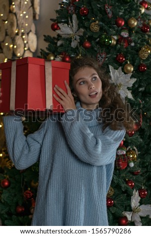 Portrait of beautiful young female model in blue sweater holding big red gift box in front of decorated Christmas tree. Concept of cosy Boxing day atmosphere. 