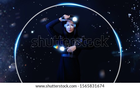 Beautiful woman in futuristic dress over dark magic background. Gamer girl in glasses of virtual reality with controllers in hands. Augmented reality, game, hobby concept. VR. Blue neon light.