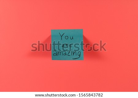 You are amazing. Blue sticky note with inspirational quote on neon pink background. Handwritten positive reminder/advice. Concept for confidence, courage and motivation. Sign of moral support.