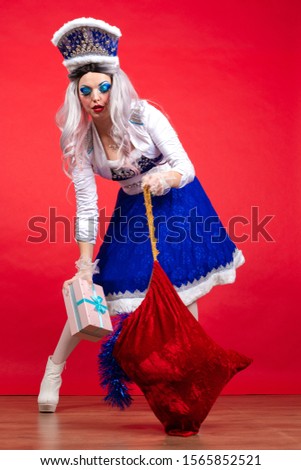 Snow Maiden in an elegant bright blue suit with a crown on his head and gifts in a bag and box posing on a red background