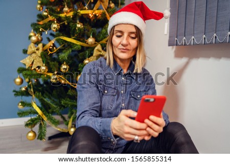 Woman female sitting at home in front of the christmas tree taking photos selfies using smart mobile phone video chat family santa hat
