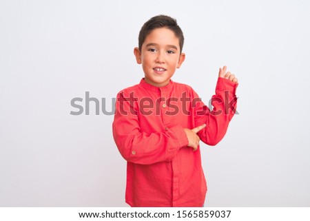 Beautiful kid boy wearing elegant red shirt standing over isolated white background with a big smile on face, pointing with hand and finger to the side looking at the camera.