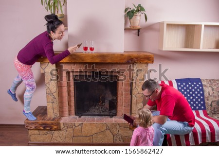 Father, mother and little daughter gathered around the fireplace in the evening. Mom takes pictures as dad plays with the baby. A family idyll.