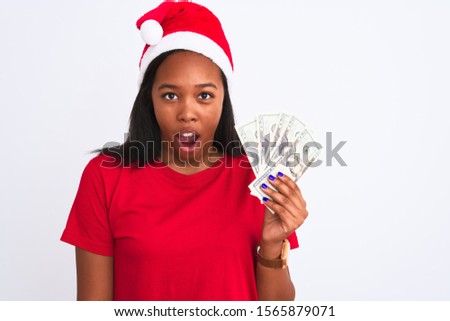 Young african american woman wearing christmas hat and holding bunch of dollars banknotes scared in shock with a surprise face, afraid and excited with fear expression
