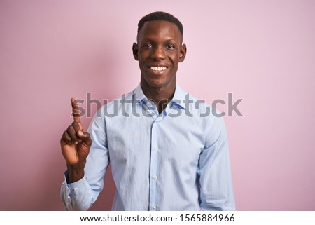 African american man wearing blue elegant shirt standing over isolated pink background showing and pointing up with finger number one while smiling confident and happy.