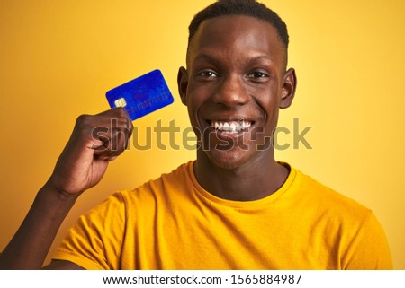 African american customer man holding credit card standing over isolated yellow background with a happy face standing and smiling with a confident smile showing teeth