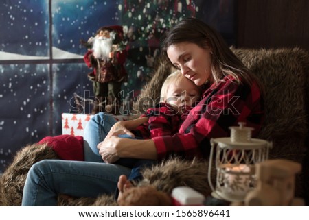 Beautiful young mother, hugging her toddler boy, sitting in cozy chair on Christmas, decoration around her
