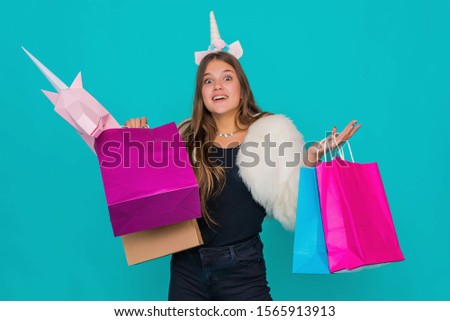 Portrait of a smiling pretty girl with shopping bags over blue background. Black friday concept. Kawaii teenage girl. Cute young woman with unicorn horn buying pink paper unicorn.