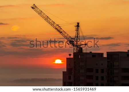 Tower crane on a construction site at sunrise with clouds. Unfinished high-rise building with luxury apartments in the city against the background of a forest in fog. Cityscape