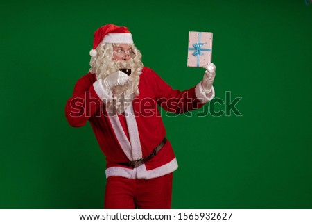 Emotional male actor in a costume of Santa Claus holds one gift box and a glass of wine in his hands and poses on a green chroma background