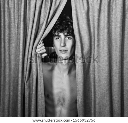 black and white photography. young handsome Italian model boy with dark curly hair posing for editorial shooting hiding behind curtains