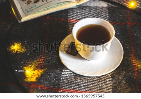 A cup of hot black tea with chocolate on a saucer on a warm and cozy plaid with Christmas lights
