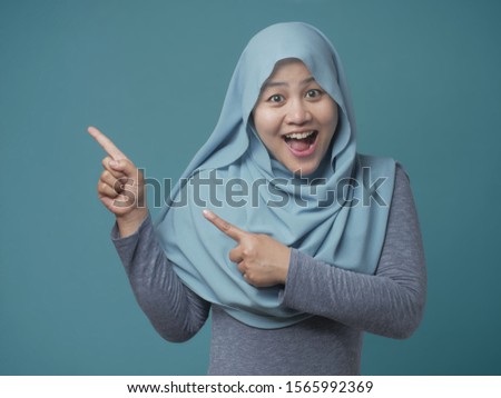 Portrait of attractive beautiful muslim woman wearing hijab  smiling and pointing to the side with copy space, against blue background
