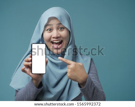 Portrait of Asian muslim woman looking at camera smiling, showing and pointing her smart phone, phone mock up, against blue background