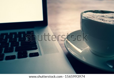 A cup of cappuccino coffee with laptop white screen on table. Royalty high quality free stock photo image of coffee cup with laptop for working in a coffee shop, typing with blank screen black color