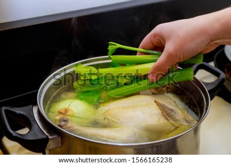 Hand puts celery in soup in the pot with whole chicken.