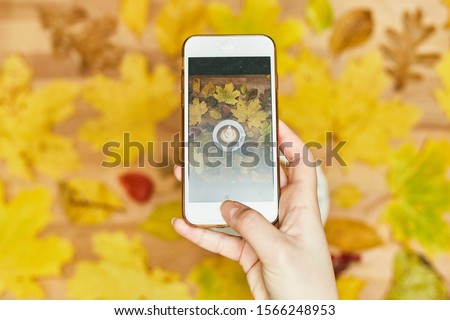 A girl photographs an autumn flatlay on her phone. Autumn flat lay composition with dry leaves wreath frame and coffee latte cup on wooden background