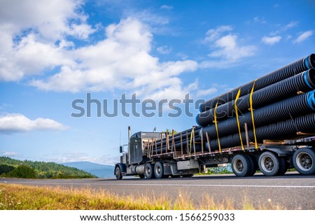 Big rig powerful professional industrial bonnet semi truck for long haul delivery commercial plastic pipes going with step down semi trailer on the summer road with green forest trees on the sides Royalty-Free Stock Photo #1566259033