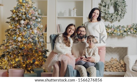Cute European family of four posing on camera for a Christmas photo in the living room of his house in an armchair near the Christmas tree and fireplace