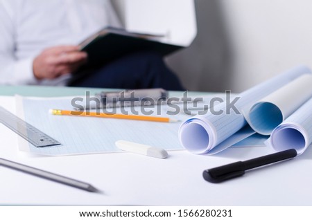 Draftsman at work, analyzing documents for drafting. Closeup of graph papers, rulers and other tools on the desk, engineer sitting on the background
