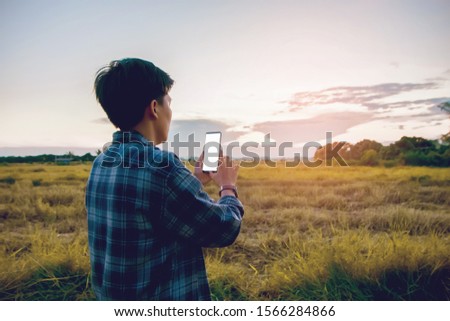 Half Back view, Smiling Asian man wearing plaid shirts and vintage eyeglasses are using a smartphone, Screen white space mock-up in evening with sunset outdoor. Young people working on mobile devices