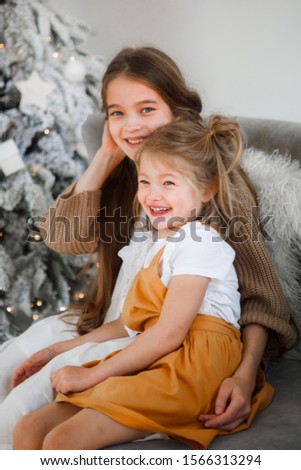Little sisters are sitting and hugging on the couch on Christmas morning.