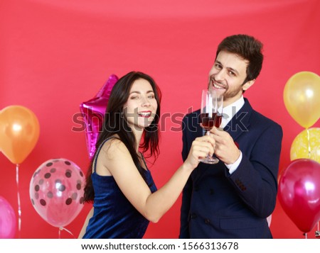 Portrait of smart man and beautiful woman holding and drinking glass of wine isolated on red background with colorful balloons - Christmas Happy New Year and Valentines party concept